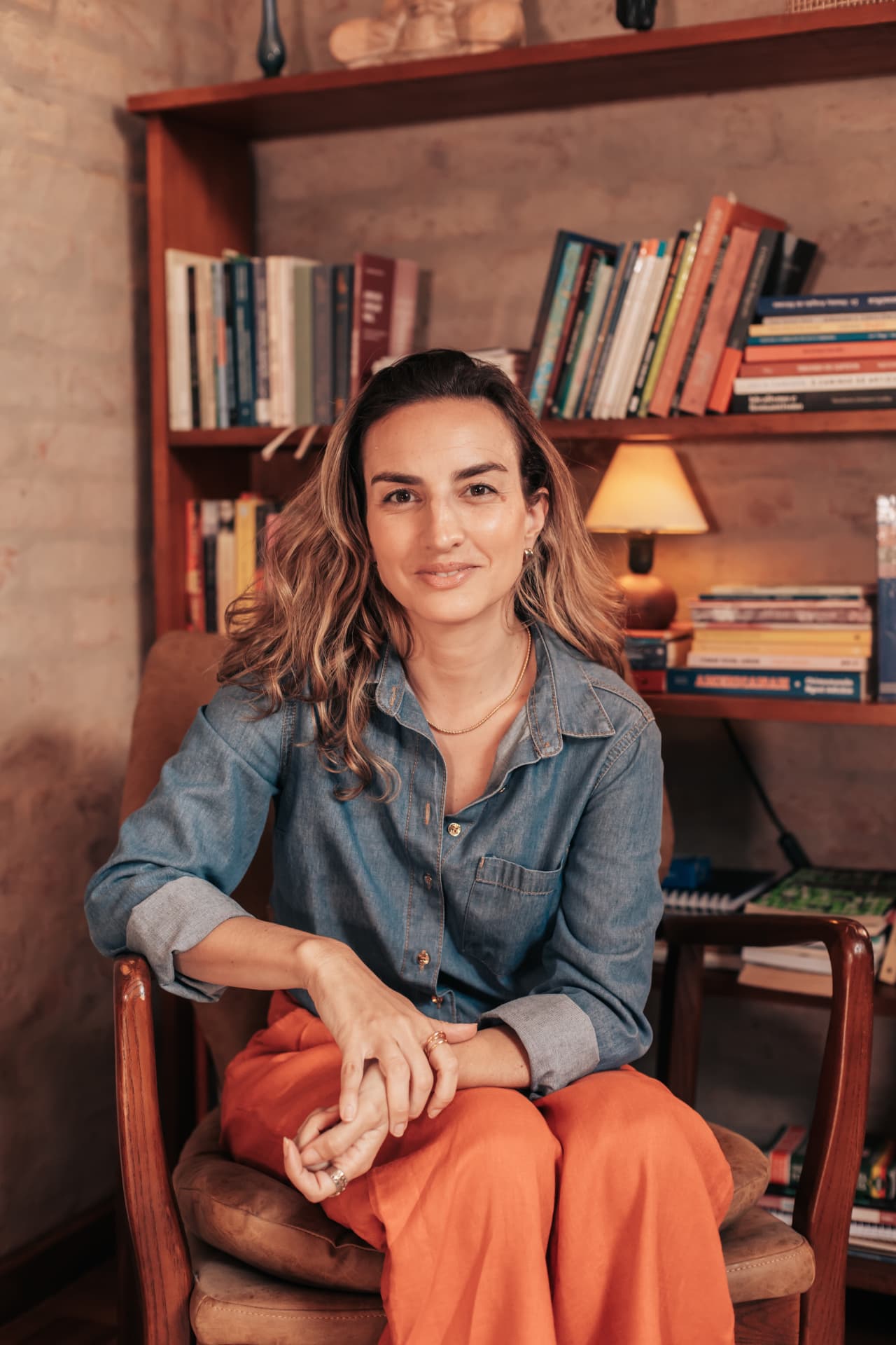 Retrato de Debora Garcia de camisa jeans e calça laranja, sentada em poltrona